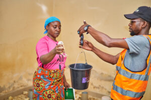 This image shows a moment of hands-on fieldwork between a Pullus extension officer and a poultry farmer inside a poultry pen. The Pullus extension officer, dressed in a reflective orange vest and black cap, is holding a portable digital scale with a black bucket suspended from it. Opposite him, the poultry farmer wearing a bright pink shirt, patterned wrapper, and a blue headscarf is holding a day-old chick, preparing to place it in the bucket for weighing.